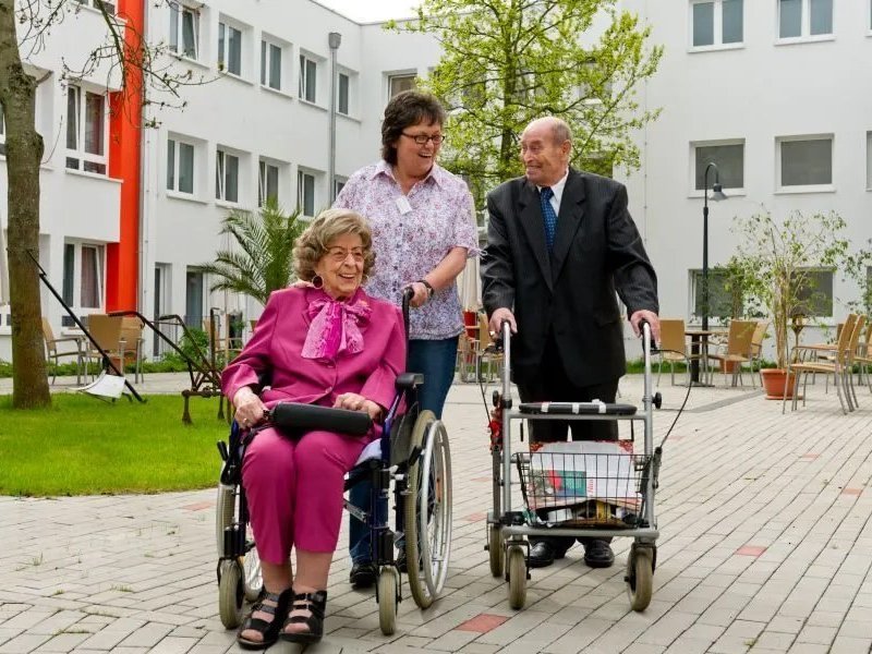 Eine ältere Frau im Rollstuhl in einem dunkelpinken Kostüm wird von einer Altenpflegerin über den Hof des Altenheims geschoben. Sie werden von einem älteren Herrn mit Rollator begleitet. Alle drei unterhalten sich und lächeln freundlich.