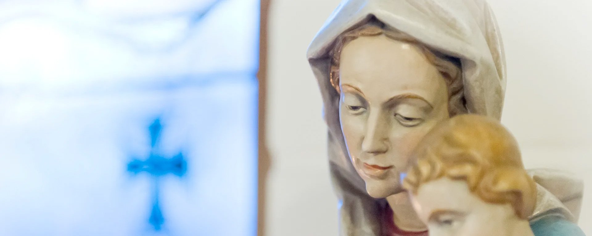 Der Kopf einer Marienstatue mit Jesuskind. Im Hintergrund ein blaues Kreuz in einem Fenster.