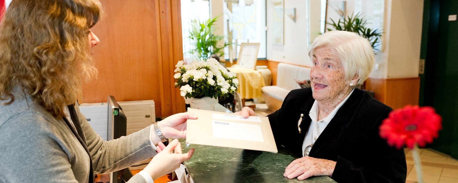 An einer Rezeption übergibt eine ältere Dame einen Umschlag an die Rezeptionistin.