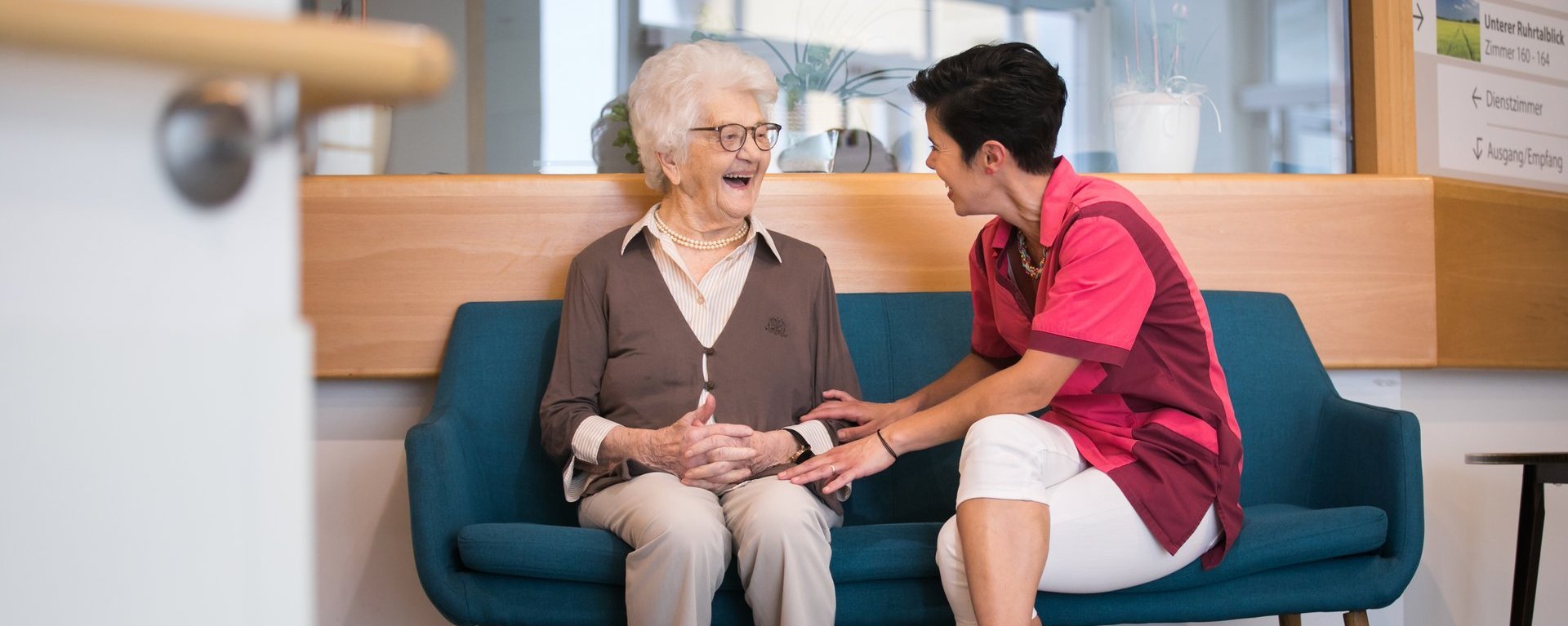 Eine Pflegerin und eine Bewohnerin sitzen auf einem Sofa, unterhalten sich und lachen