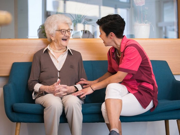 Eine Pflegerin und eine Bewohnerin sitzen auf einem Sofa, unterhalten sich und lachen