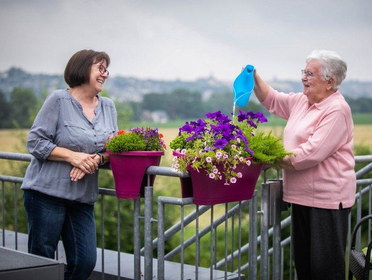 Zwei Personen stehen auf einem Balkon und die Bewohnerin des Altenheims gießt mit einer Gießkanne Blumen.