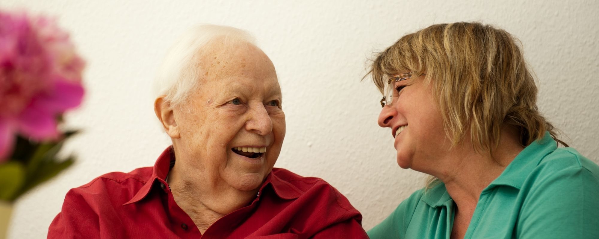 Eine ältere Dame und eine Altenpflegerin sitzen nebeneinander und lachen zusammen.