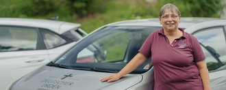 Eine Mitarbeiterin des ambulanten Pflegedienstes lehnt an ihrem Dienstfahrzeug und lächtelt in die Kamera.