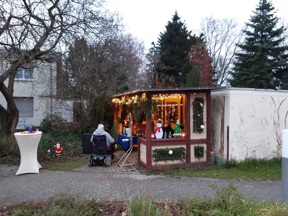 In einem herbstlichen Park steht ein weihnachtlicher Stand mit Lichterketten. Ein Mann im Rollstuhl bewegt sich darauf zu. 